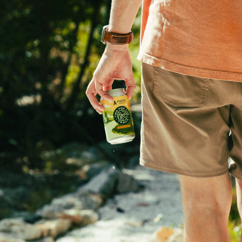 A close-up of a person's hand holding a chilled can of Two For The Trails Milestone Edition IPA while walking on a sunlit trail.