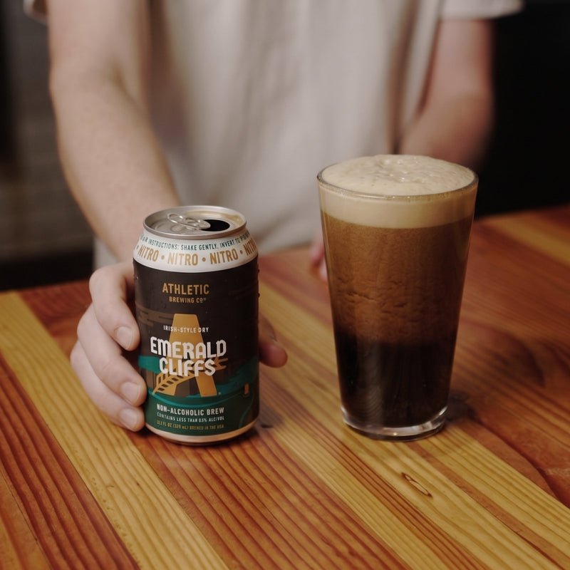 Athletic Brewing Emerald Cliffs Irish-Style Dry non-alcoholic nitro brew can beside a glass of dark beer with a thick, creamy foam head on a wooden table.