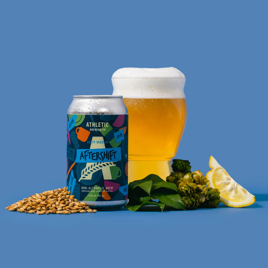 Can of Aftershift non-alcoholic brew next to a glass of non-alcoholic brew next to lemon, hops, and yuzu leaf