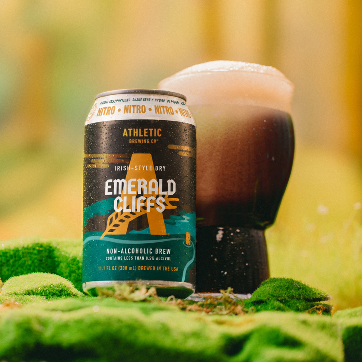 Athletic Brewing Emerald Cliffs non-alcoholic nitro brew can beside a glass of dark beer with a creamy foam head in a mossy outdoor setting