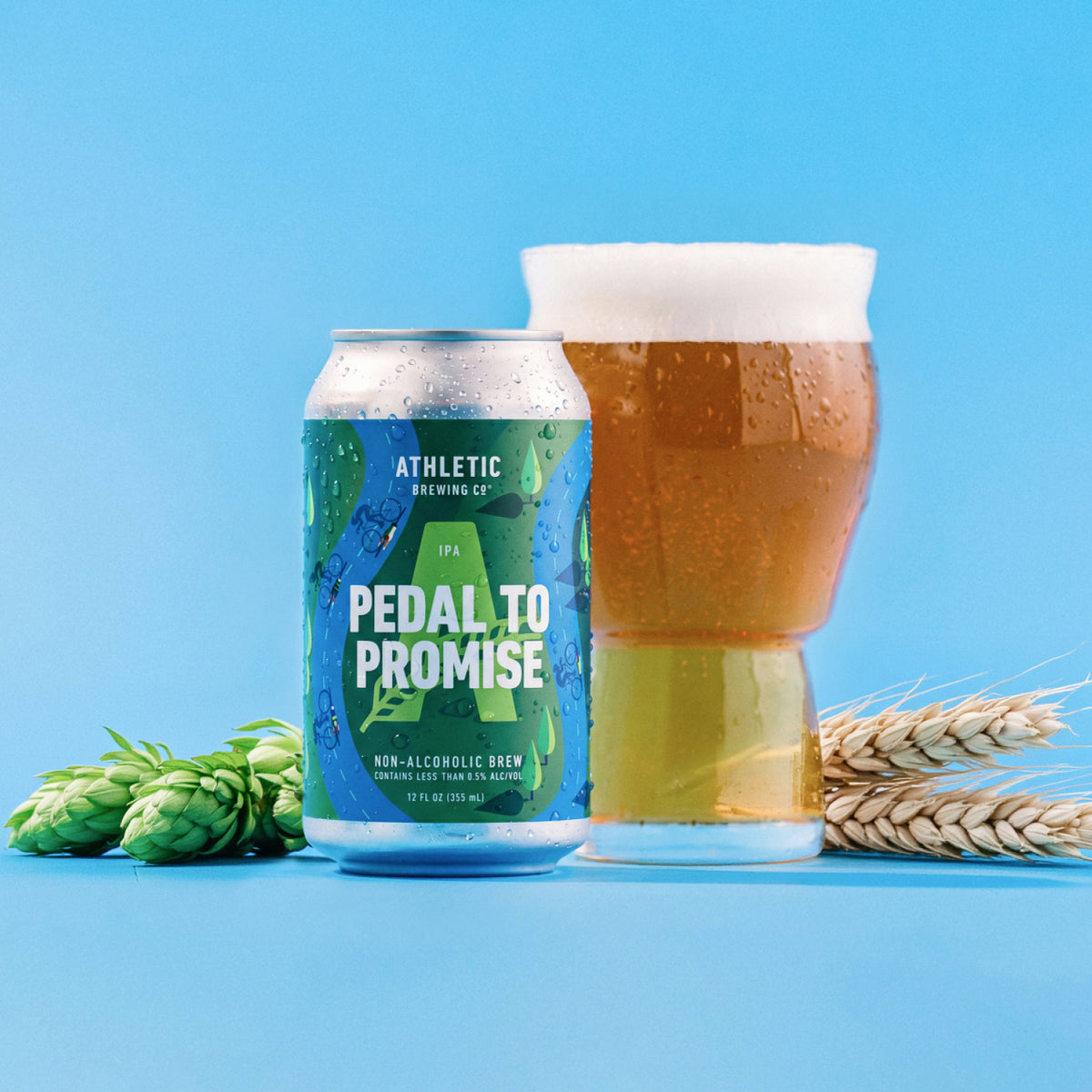 Athletic Brewing Pedal to Promise non-alcoholic IPA can beside a glass of golden beer topped with foam. Fresh hops and wheat frame the can and glass against a bright blue background.