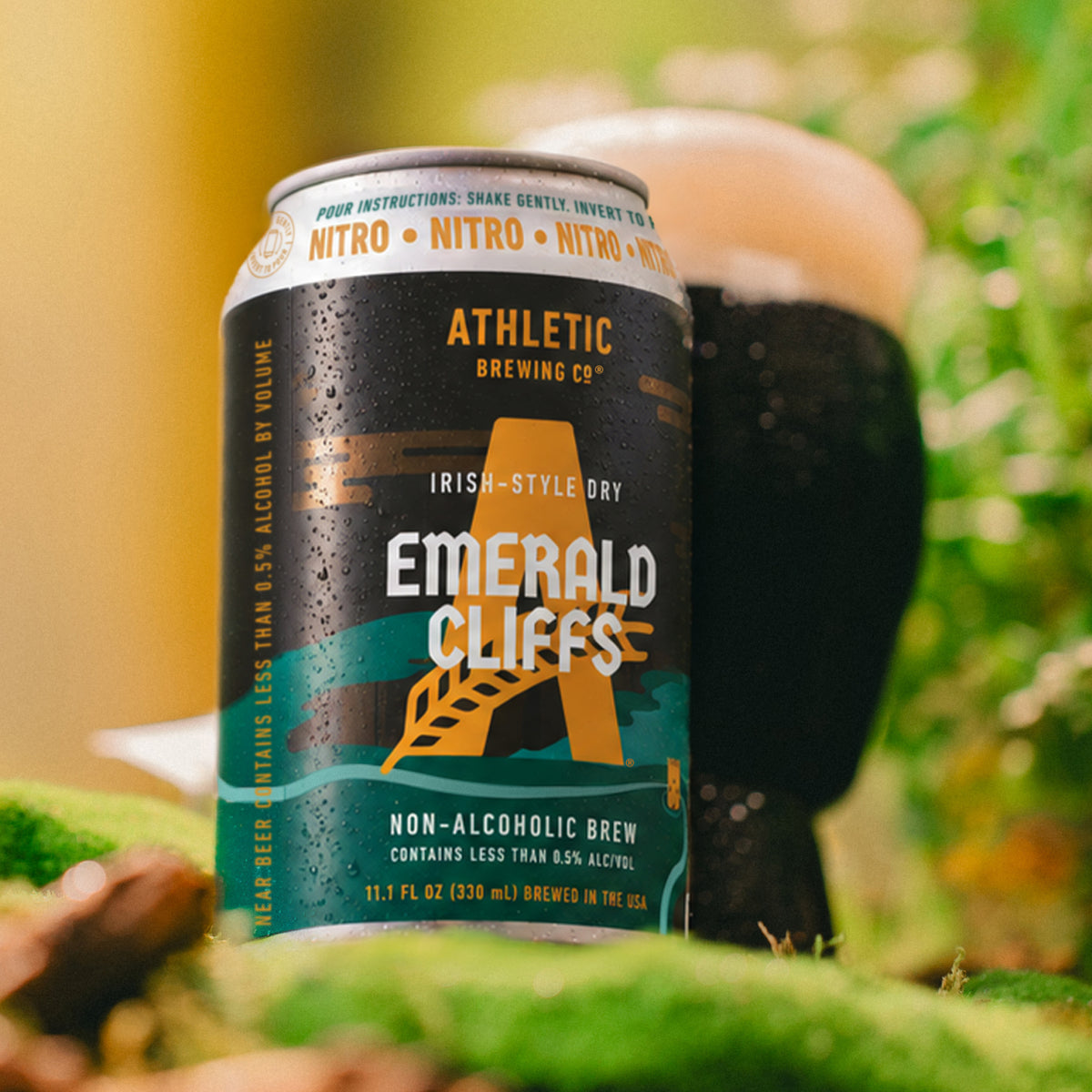 Chilled Athletic Brewing Emerald Cliffs non-alcoholic nitro brew can with a dark, foamy pour in the background, surrounded by lush green scenery.