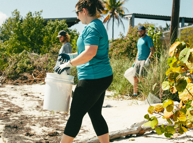 A team of volunteers in teal shirts participating in a beach cleanup, with one person carrying a white collection bucket.